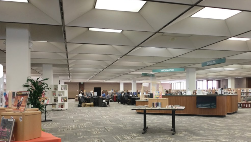 A spacious library interior with bookshelves, study areas, and people working at desks under bright, geometric ceiling lights.