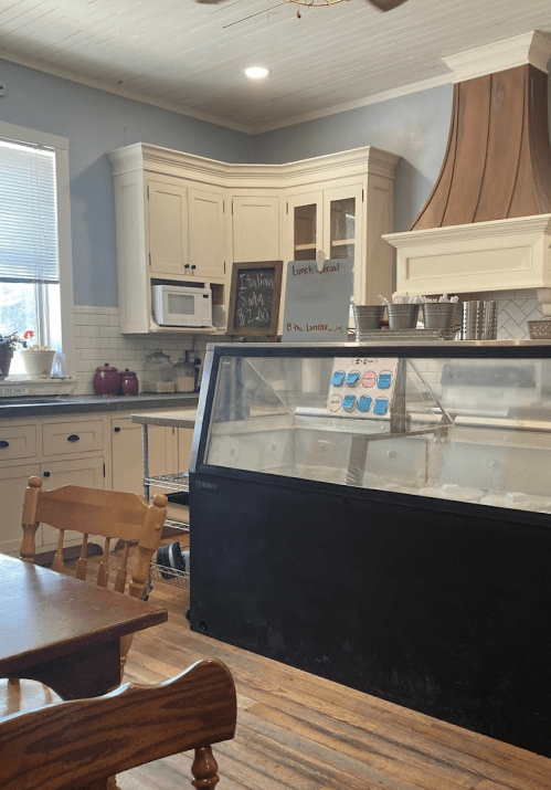 A cozy café interior featuring a glass display case, wooden tables, and a kitchen area with cabinets and appliances.