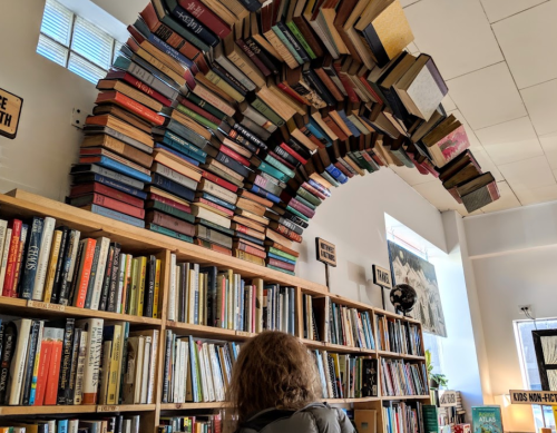 A person gazes up at a unique arch of books suspended above a bookshelf in a cozy library setting.