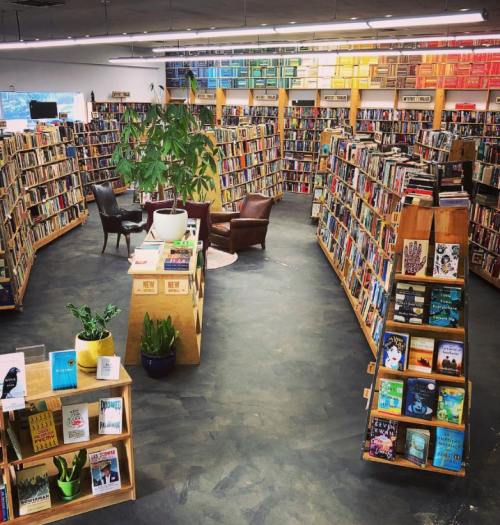 A cozy bookstore with tall shelves filled with books, a seating area, and colorful artwork on the walls.