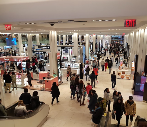 A busy department store with shoppers browsing beauty products and cosmetics, featuring bright displays and escalators.