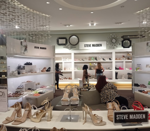 A shoe store display featuring various styles, with customers browsing and trying on shoes. Steve Madden branding visible.