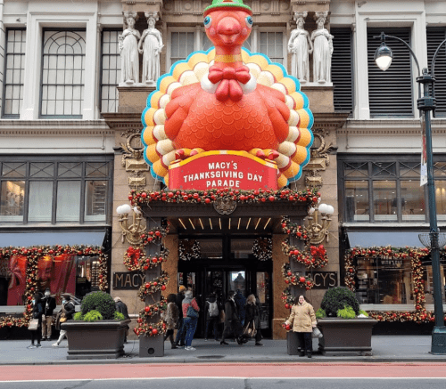 Entrance of Macy's decorated for Thanksgiving, featuring a large turkey and festive garlands. Crowds gather outside.