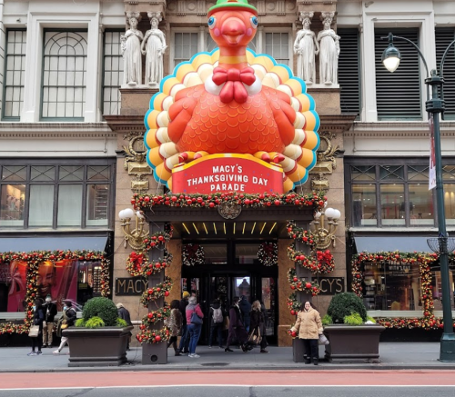 Entrance of Macy's decorated for Thanksgiving, featuring a large turkey and festive garlands. Crowds gather outside.