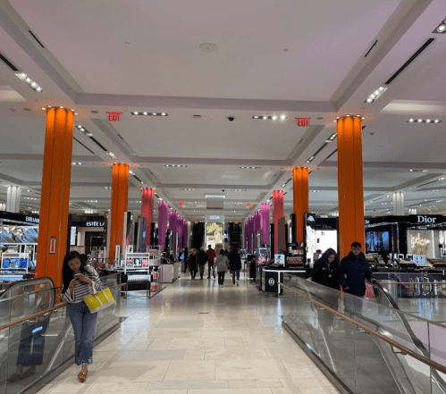 A spacious shopping mall interior with colorful lighting, featuring various beauty and fashion stores. Shoppers walk through.