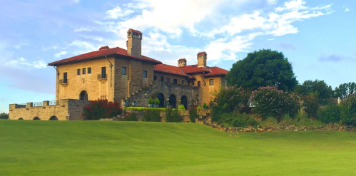 A large, historic stone mansion with a red tile roof, surrounded by green grass and trees under a blue sky.
