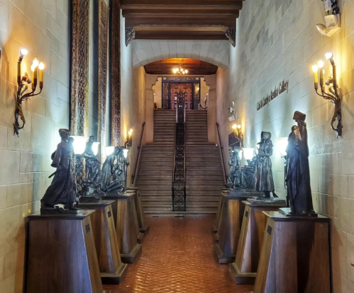 A hallway with statues on pedestals, leading to a staircase and ornate lighting on the walls.