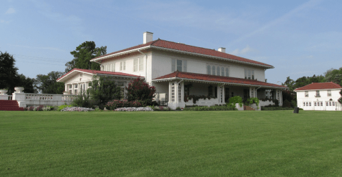 A large white house with a red roof, surrounded by green grass and colorful flower beds under a clear blue sky.
