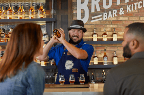 A bartender in a blue shirt and hat shakes a cocktail while two patrons watch at a bar with whiskey bottles in the background.