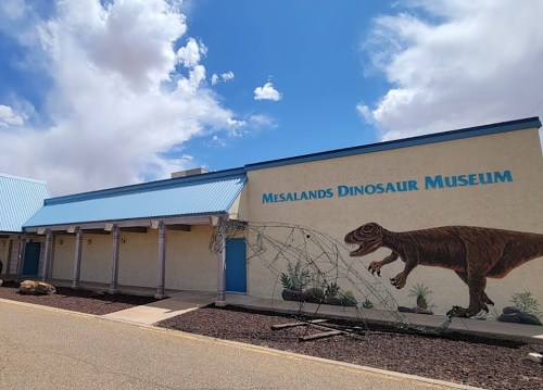 Exterior of the Mesalands Dinosaur Museum featuring a dinosaur mural and a blue roof under a cloudy sky.
