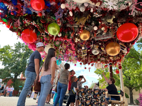 A festive display of colorful ornaments hangs overhead as people admire the decorations in a lively outdoor setting.