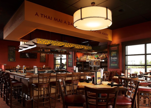 Interior of a Thai restaurant featuring a bar, wooden tables, and warm lighting. Walls adorned with decorative text.