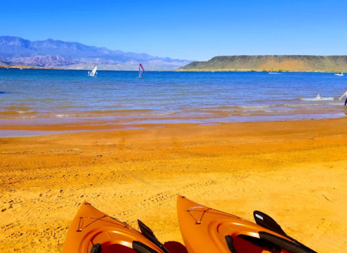 Two orange kayaks on a sandy beach with calm blue water and distant mountains under a clear blue sky.