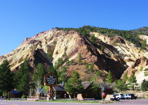 A colorful mountain landscape with a rustic lodge at the base, surrounded by pine trees and clear blue skies.