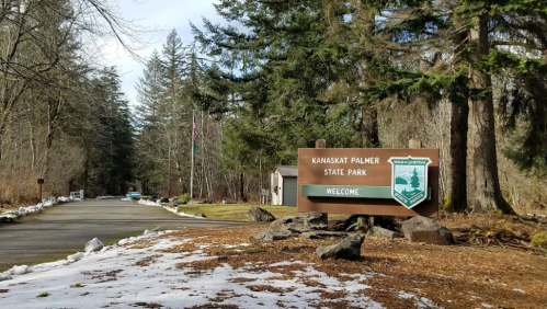Sign for Kanaskat Palmer State Park, surrounded by trees and a snowy path leading into the park.