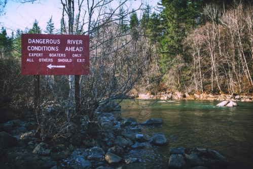 A red warning sign by a river reads "Dangerous River Conditions Ahead" with a view of trees and rocky riverbank.