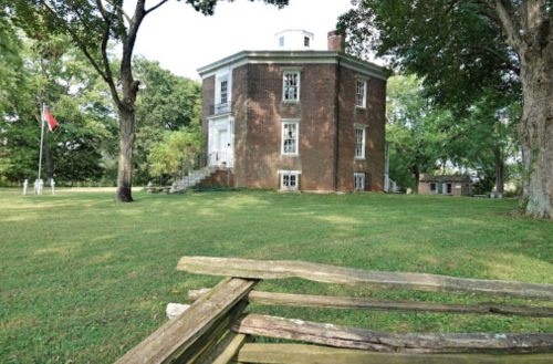 Historic brick building surrounded by trees and grass, with a flagpole in the foreground and a staircase leading up.