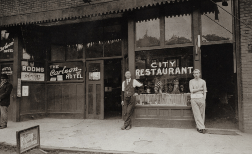 Historic black-and-white photo of a restaurant and hotel storefront, with two men standing outside.