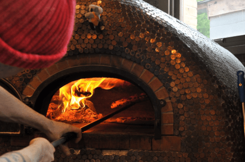 A person in a red beanie uses a tool to place a pizza into a wood-fired oven adorned with copper coins.