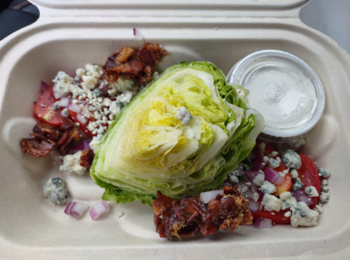 A salad with a wedge of lettuce, bacon bits, tomatoes, red onions, blue cheese, and a dressing cup in a takeout container.