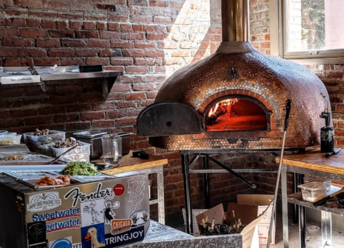 A rustic kitchen with a brick pizza oven, surrounded by food prep stations and various ingredients on the counter.