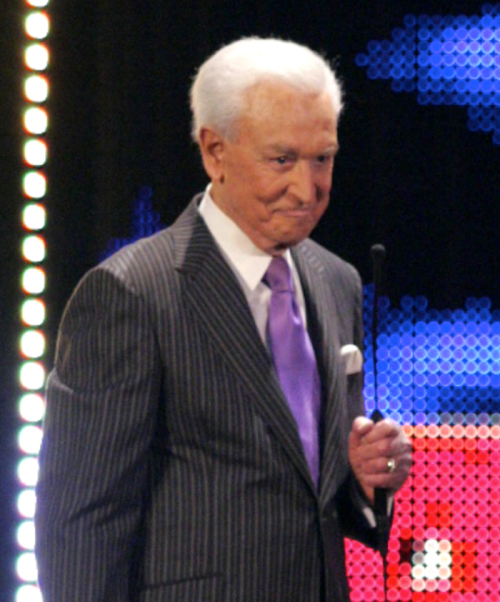 A man in a pinstripe suit and purple tie stands on stage, holding a microphone, looking thoughtful.
