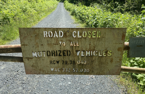 A rusty sign on a gate reads "Road Closed to All Motorized Vehicles" with legal codes below. Lush greenery surrounds the path.