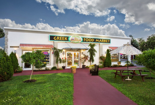 Exterior of a Greek food market with a colorful sign, surrounded by greenery and a picnic area under a large umbrella.