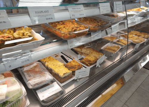A display case filled with various prepared dishes, including moussaka, eggplant, and pastries.