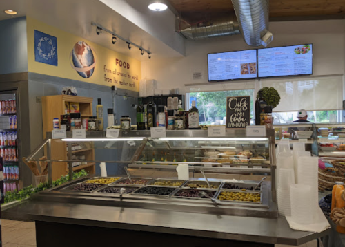A bright café interior featuring a food display with various olives and condiments, menus on the wall, and a welcoming atmosphere.