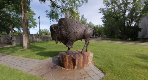 A bronze statue of a bison stands on a stone pedestal, surrounded by green grass and trees.