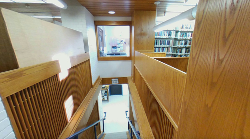A wooden staircase leads down to a library space, with shelves visible in the background and natural light coming through a window.