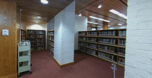 A cozy library interior with bookshelves lining the walls and a cart for books in the foreground.