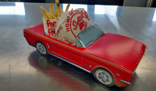A red toy car with a bag of fries on top, labeled "Home of the Best Fries," on a shiny metal surface.