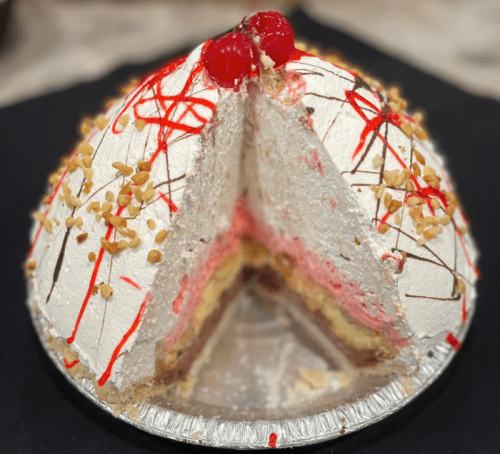 A dome-shaped cake with layers of cream, cherries on top, and a slice revealing colorful layers inside.