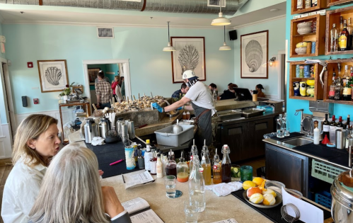 A busy restaurant bar with patrons, a bartender preparing oysters, and a variety of drinks and garnishes on the counter.