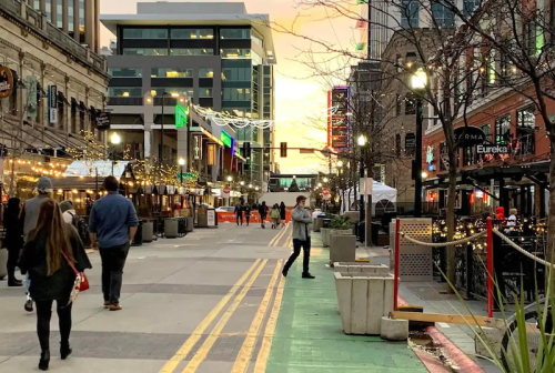 A bustling street at sunset, lined with shops and festive lights, with people walking and enjoying the atmosphere.