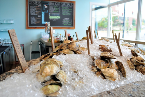 A display of oysters on ice with wooden markers, set in a bright restaurant with a chalkboard menu in the background.