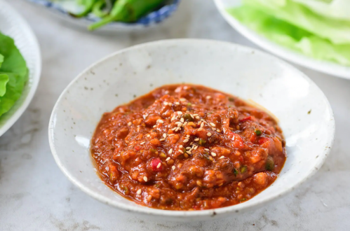 A bowl of spicy red sauce topped with sesame seeds, surrounded by fresh lettuce and green peppers.