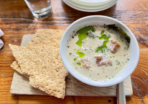 A bowl of creamy soup with green oil and croutons, served with crispy flatbread on a wooden board.