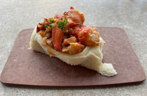 A lobster roll topped with chives, served on a wooden board.