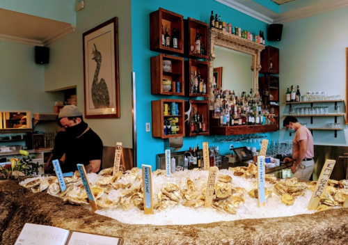 A bar with a display of oysters on ice, surrounded by bottles and a bartender in a mask, with a colorful wall behind.