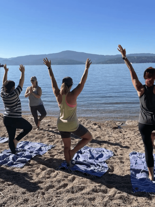 Four women practicing yoga on a beach, with arms raised, against a backdrop of water and mountains.