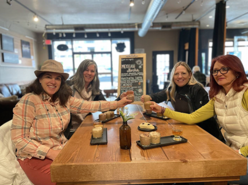 Four women sitting at a wooden table in a café, smiling and toasting with drinks, surrounded by a cozy atmosphere.