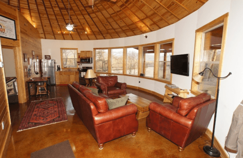 Cozy living room with leather sofas, wooden ceiling, large windows, and a rustic decor.