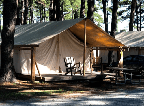 A canvas tent on a wooden platform surrounded by trees, featuring an Adirondack chair and a picnic table nearby.