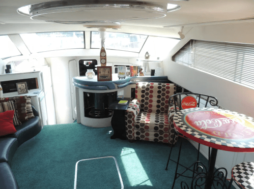 Cozy interior of a boat featuring a colorful seating area, a small table, and a kitchenette with large windows.