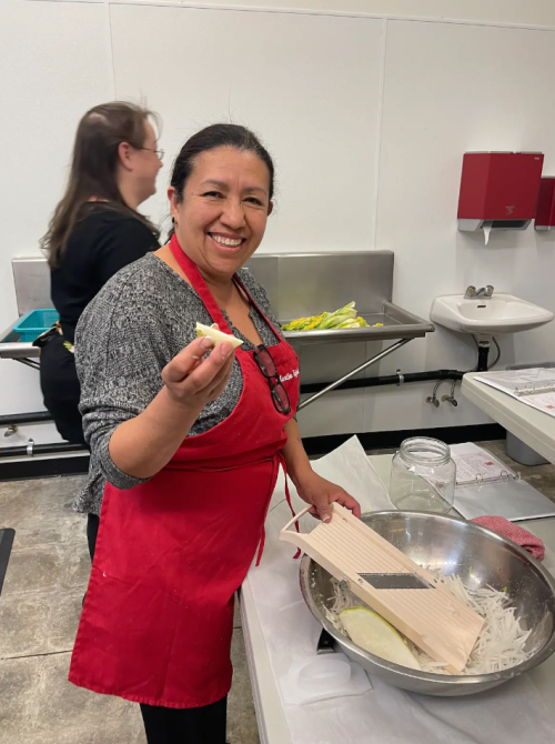 A smiling woman in a red apron holds a piece of food while preparing ingredients in a kitchen setting.