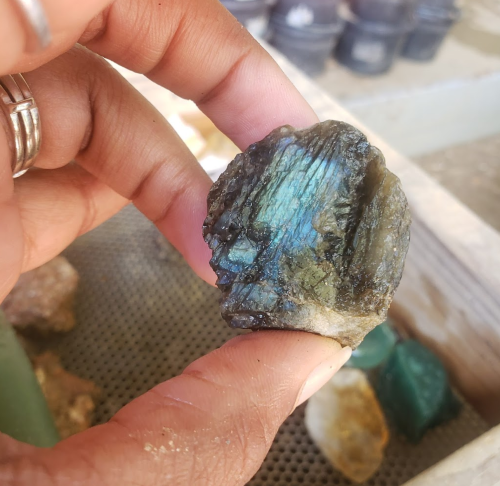 A hand holding a shiny, iridescent mineral with green and blue hues against a blurred background of rocks.