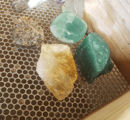 A collection of colorful crystals and stones, including yellow, green, and clear varieties, on a mesh surface.